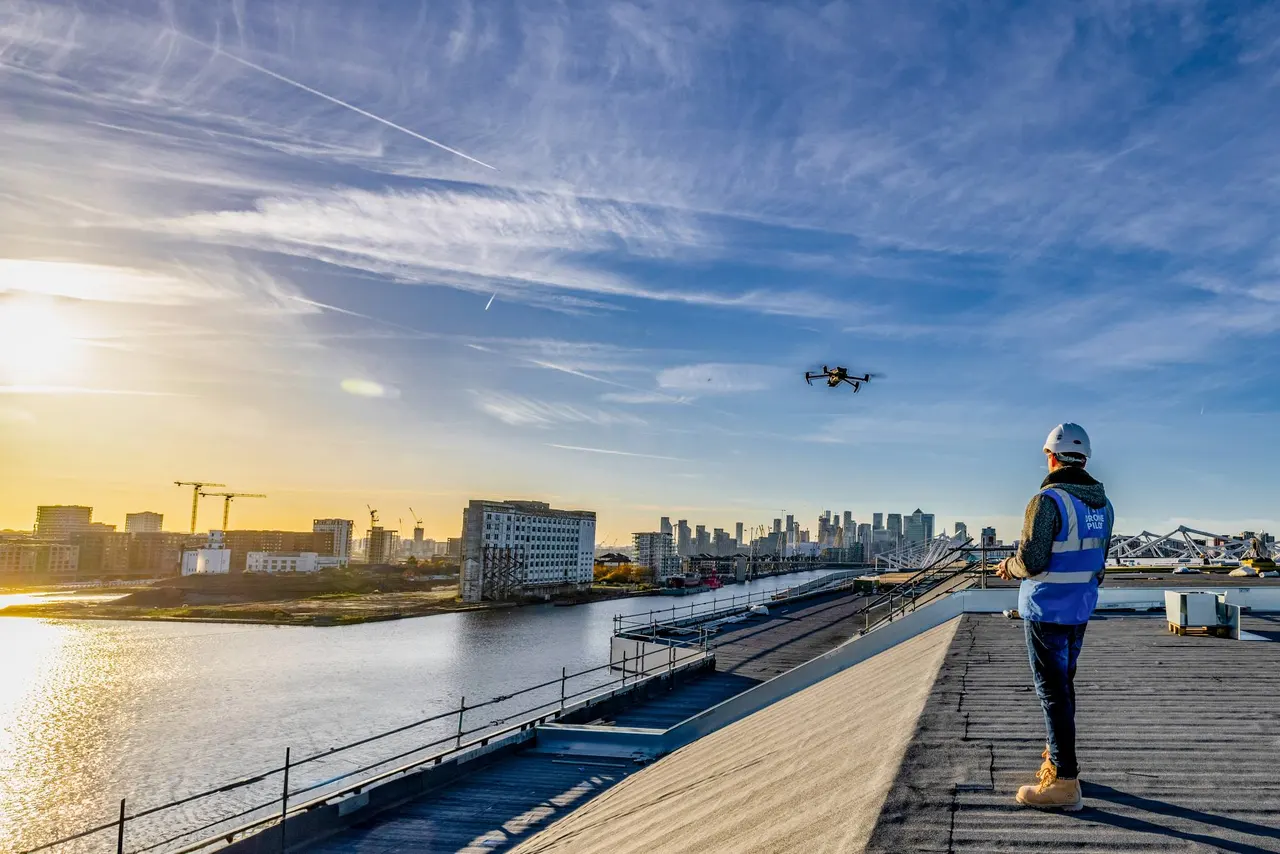 A drone pilot on a rooftop beside the Thames, with a drone against the evening sky